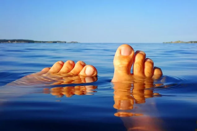Fötter från en badare sticker upp ur vattnet i en stilla skärgård på sommaren. Foto. 