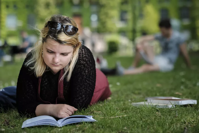 En student ligger på mage i gräset och läser framför Universitetsbiblioteket en sommardag. Foto.